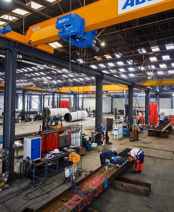 Atelier de soudage industriel et ponts roulants ABUS Ouvriers soudant une poutre d'acier dans un atelier industriel avec des ponts roulants ABUS jaunes et bleus.